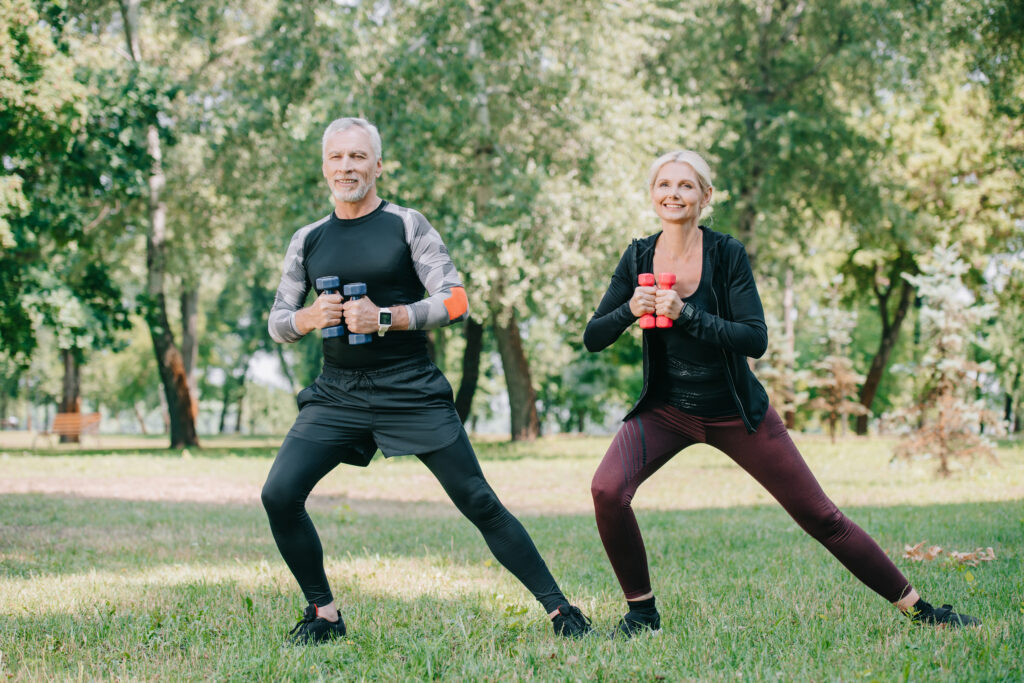 cheerful mature sportsman and sportswoman exercising with barbells in park